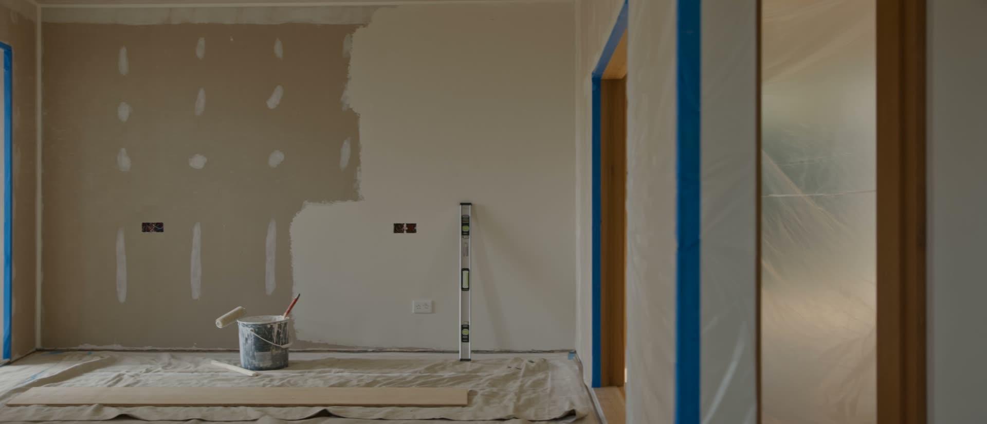 A room mid-renovation: patched plasterboard part-painted, masking tape, a paint tin and spirit level on drop sheets
